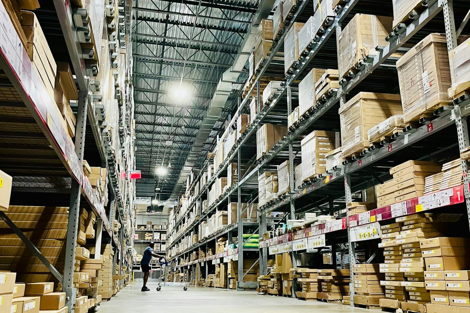 Interior of a large distribution warehouse with tall racking stacked with boxed goods and a worker moving a cart down the aisle, evoking a Mexico market-entry supply chain