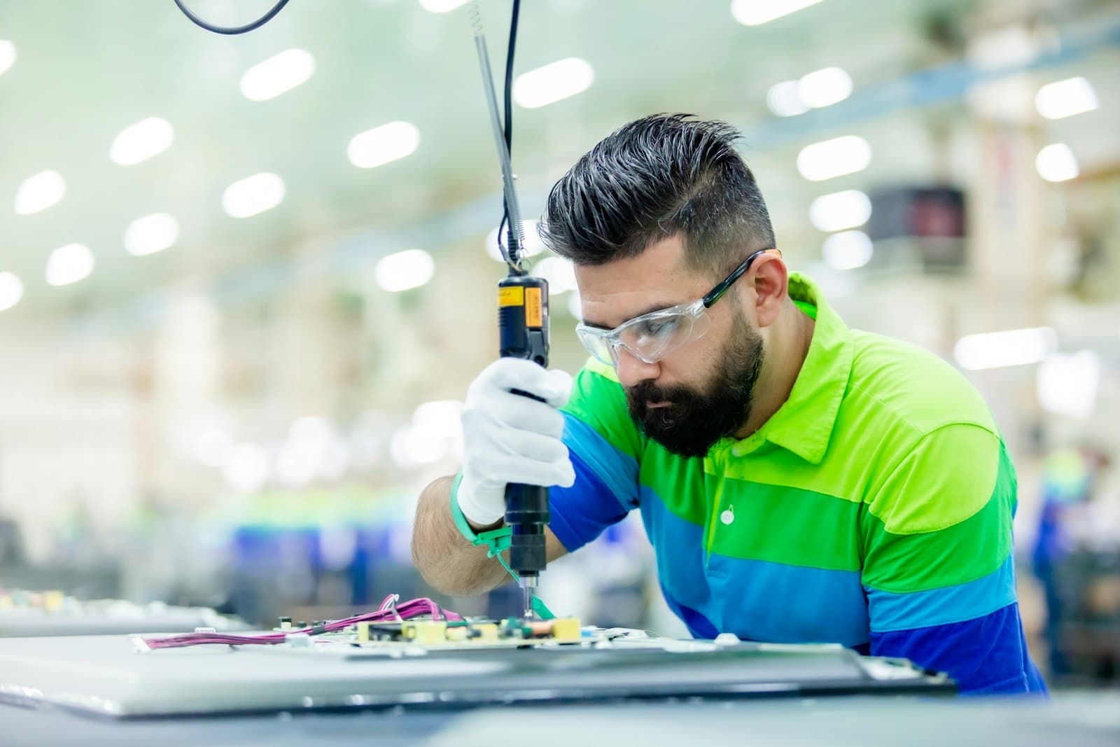A technician in safety glasses and gloves uses a torque driver to assemble an electronic circuit board on a contract manufacturing assembly line