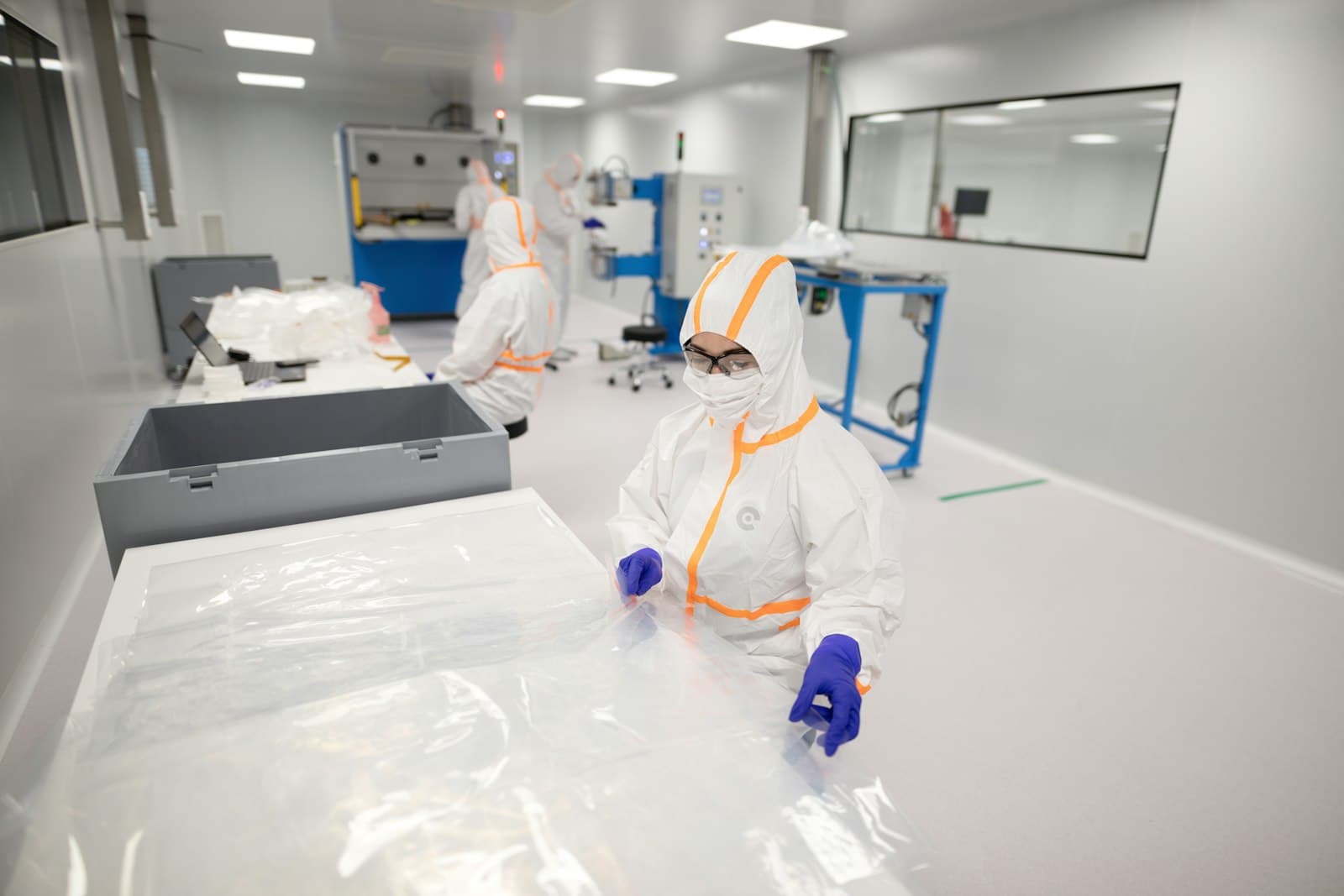 Technicians in full white cleanroom coveralls, hoods, masks, and gloves working inside a bright sterile GMP cleanroom with blue manufacturing equipment, the kind of regulated medical-device and pharmaceutical production environment Mexico entrants must certify to under COFEPRIS.