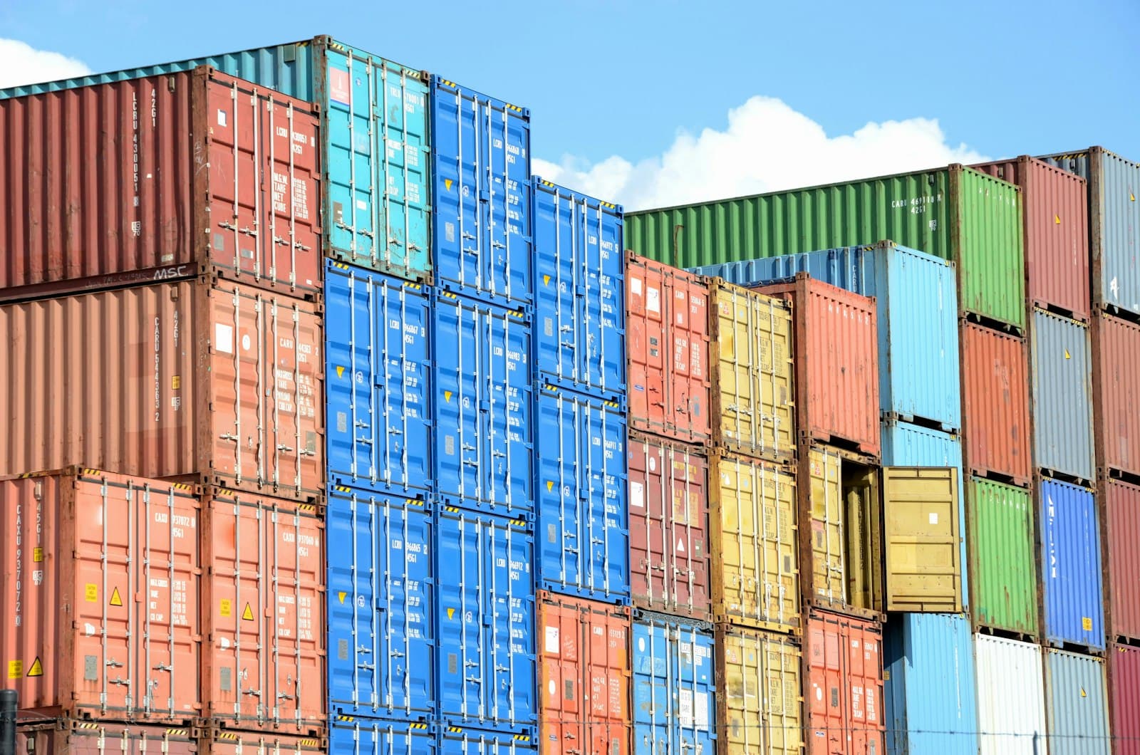 A towering wall of multicolored intermodal shipping containers stacked against a clear blue sky, evoking the total landed cost and trade comparison between Mexico and China for goods bound for the North American market