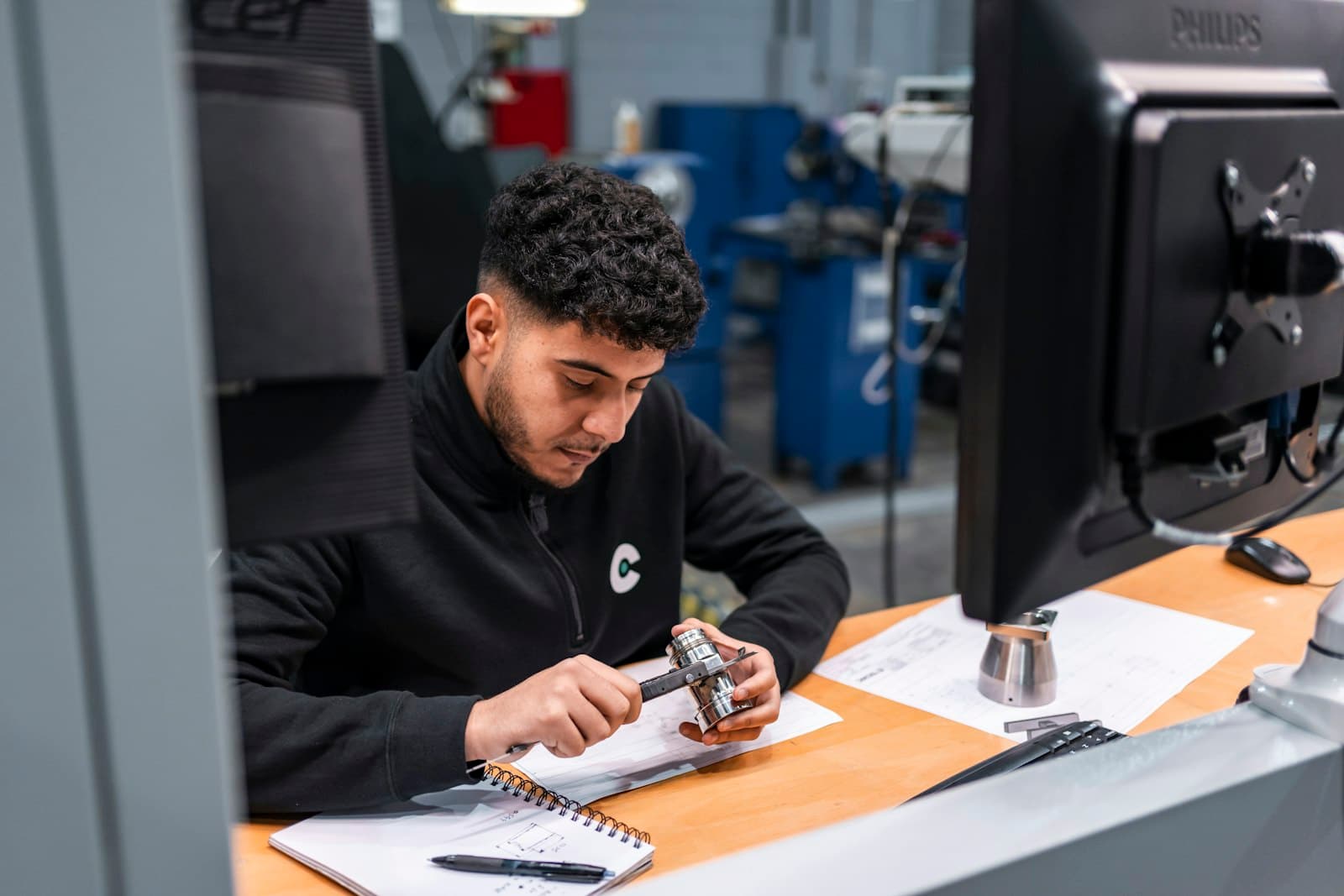 A machinist checks a turned steel part with calipers at a workstation beside the machine shop floor, the skilled hour whose cost any Mexico versus United States comparison has to price