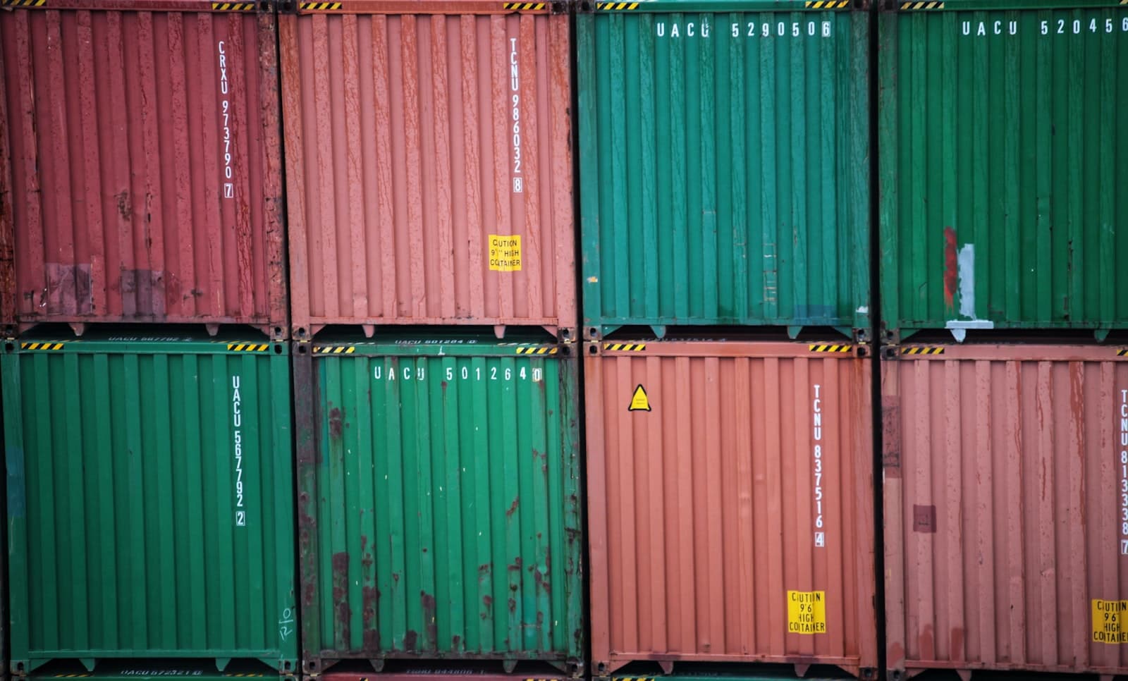 Stacks of weathered green and rust-red intermodal shipping containers at a freight terminal, evoking the North American trade flows and rules-of-origin questions at the center of the 2026 USMCA review
