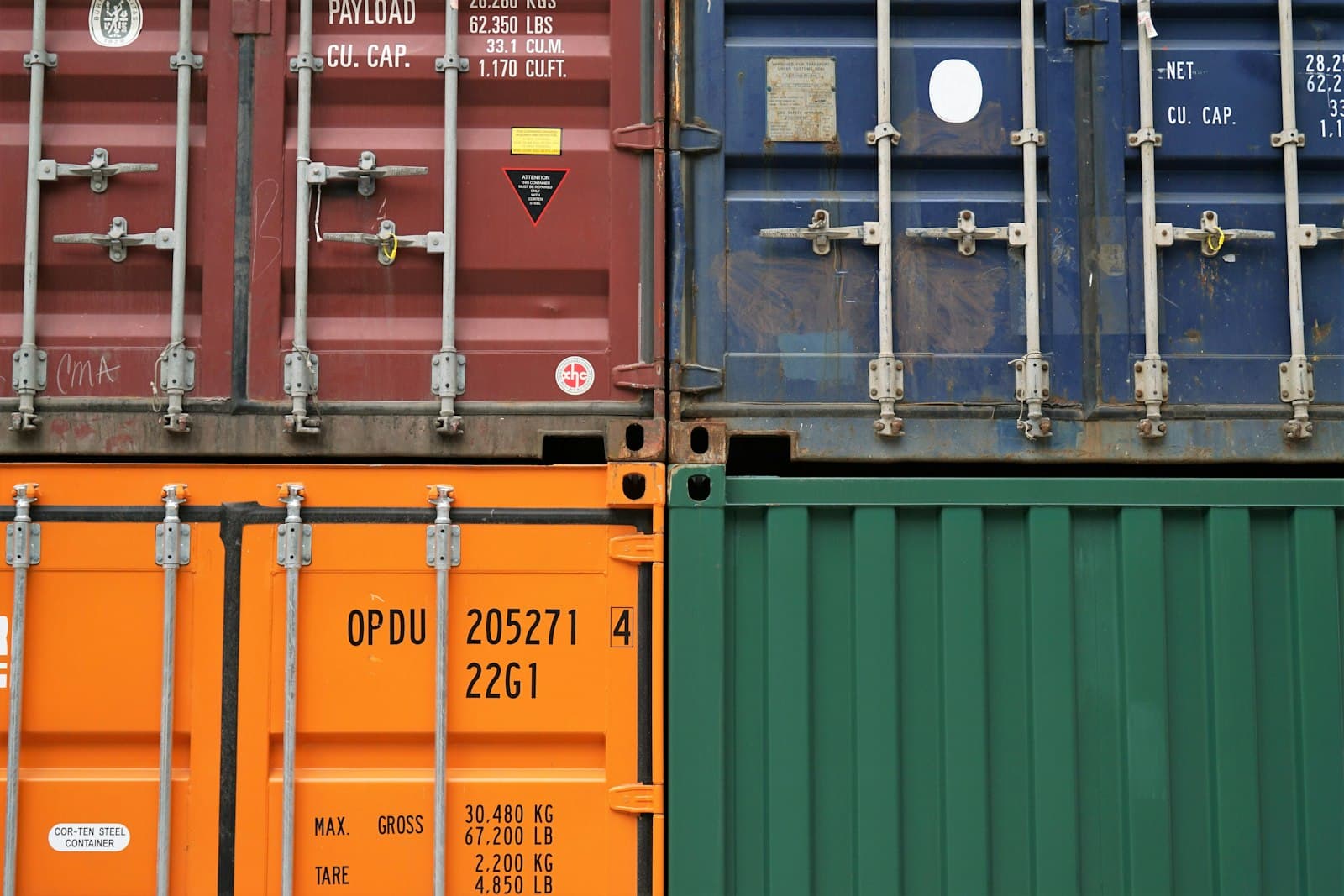 Stacked shipping containers in red, blue, orange, and green, their doors covered in payload, capacity, and country-of-origin markings and container codes, at a Mexican cargo terminal.