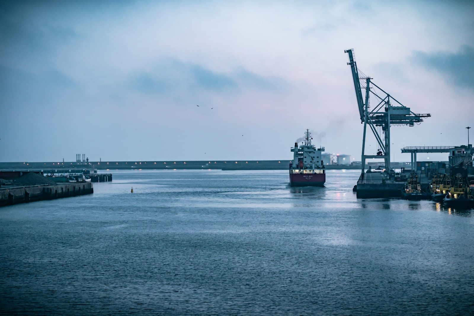 A cargo ship enters a working North American port at dusk past a gantry crane, evoking the treaty-backed US market access Mexico still holds after the July 2026 Section 338 tariffs on Canada