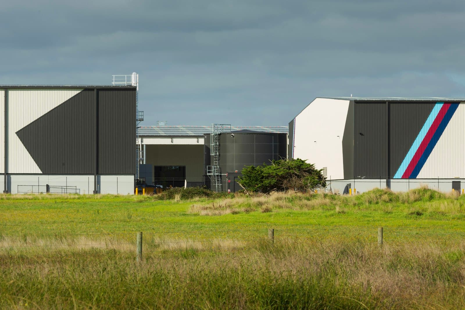 Modern industrial park buildings and storage tanks set behind an open grassy field under a cloudy sky